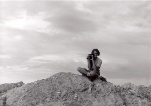 young man sitting atop rock looking through video camera